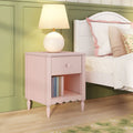 Pink nightstand with drawer and open shelf holding books, round table lamp beside white bed against pastel green paneled wall — girls' bedroom decor and bedside table idea