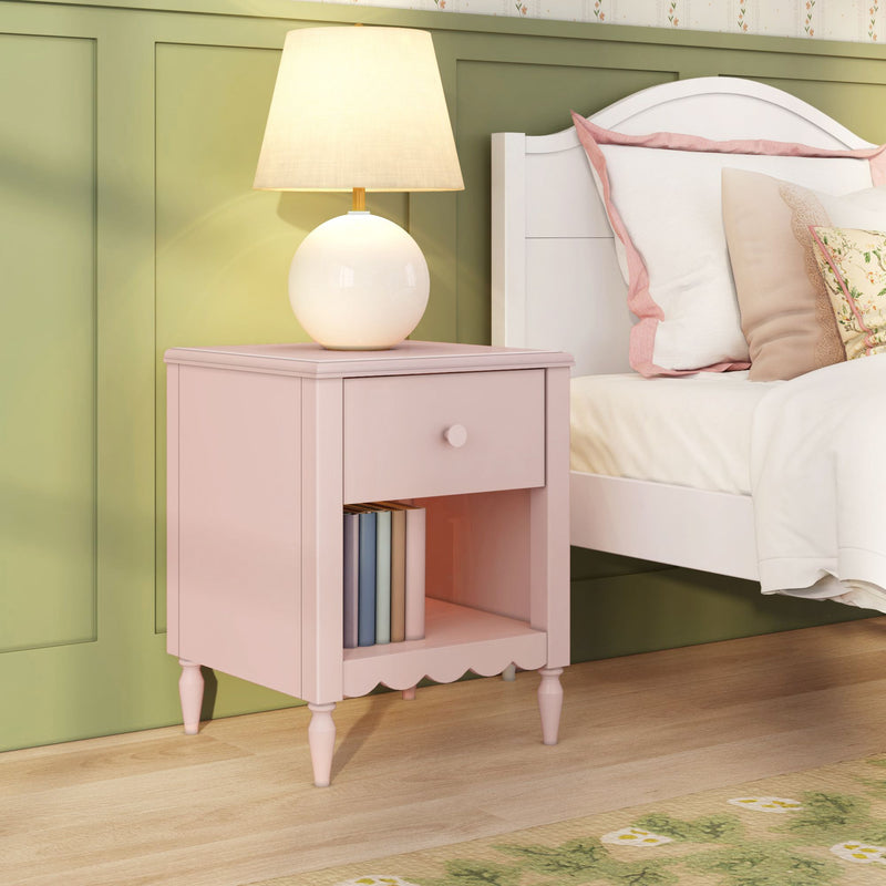 Pink nightstand with drawer and open shelf holding books, round table lamp beside white bed against pastel green paneled wall — girls' bedroom decor and bedside table idea