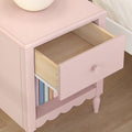 Pink wooden nightstand with round lamp, open drawer revealing light wood interior and lower shelf storing pastel books, nursery bedside table