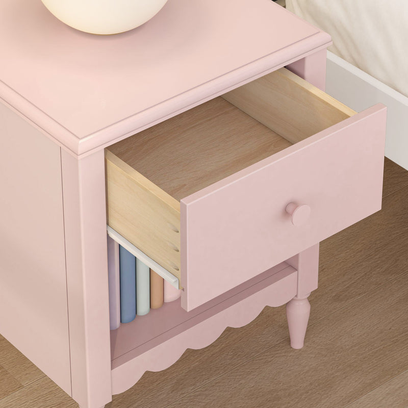 Pink wooden nightstand with round lamp, open drawer revealing light wood interior and lower shelf storing pastel books, nursery bedside table