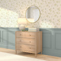 Light wood dresser with four drawers beneath round mirror, ceramic table lamp and stacked books against pastel floral wallpaper and blue wainscoting — cozy nursery or bedroom decor