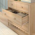 Light wood dresser with open drawer of folded clothes, wooden knobs and scalloped base — bedroom storage chest of drawers
