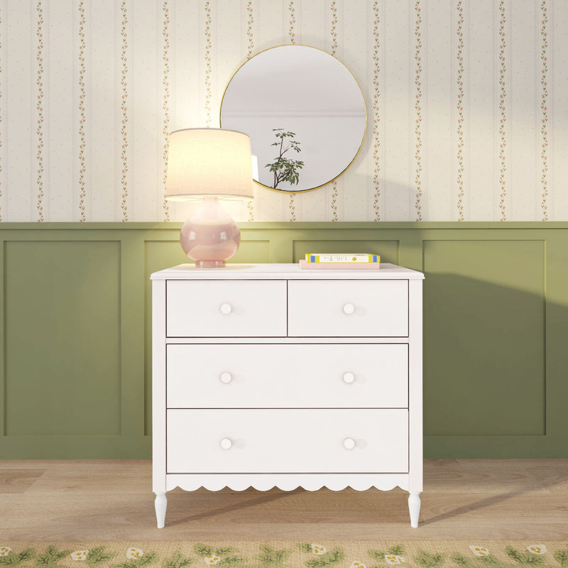 White four-drawer dresser with scalloped trim in nursery, pink lamp and round mirror, green wainscoting and floral wallpaper.