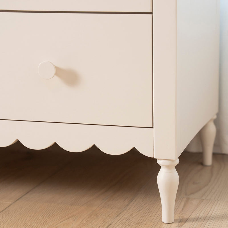 Close-up of cream painted wooden nightstand with round drawer knob, scalloped apron and turned leg on light hardwood floor
