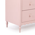 Close-up of a pink painted wooden dresser corner showing drawers with round knobs, scalloped skirt and turned spindle legs