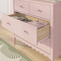 Pink nursery dresser with open drawer of folded baby clothes, round knobs and scalloped trim on light hardwood floor next to green patterned rug