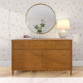 Six-drawer wooden dresser with scalloped apron topped with white ceramic table lamp, pink headphones on a book and round gold mirror reflecting a potted plant, set against delicate floral wallpaper — cozy bedroom dresser decor