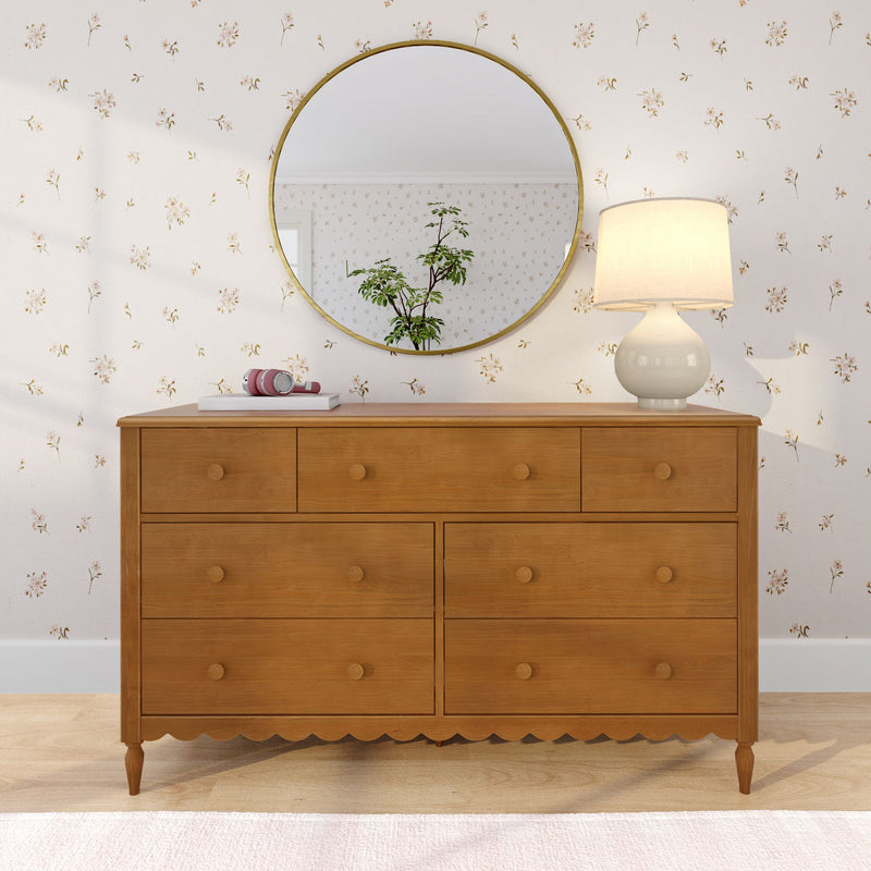 Six-drawer wooden dresser with scalloped apron topped with white ceramic table lamp, pink headphones on a book and round gold mirror reflecting a potted plant, set against delicate floral wallpaper — cozy bedroom dresser decor