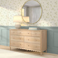 Light wood six-drawer dresser with ceramic table lamp and round gold mirror against floral wallpaper and mint wainscoting in a bright modern interior
