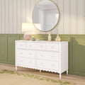 White six-drawer nursery dresser with scalloped skirt and round gold mirror, pink table lamp and wooden house decor against green wainscoting and floral wallpaper