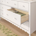White wooden nursery dresser with scalloped trim and round knobs, open drawer revealing folded baby clothes on hardwood floor and floral rug