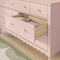 Pink wooden nursery dresser with scalloped trim and round knobs, open drawer revealing folded baby clothes on light wood floor and green patterned rug