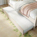 Space-saving white trundle bed pulled out from under a cream wooden twin bed with white bedding, blush pink throw and floral area rug