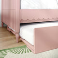 Pink scalloped bed frame with white mattress and under-bed trundle on hardwood floor with green area rug