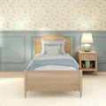 Cozy cottage bedroom with light wood twin bed, scalloped headboard, soft blue bedding, pale blue wainscoting, floral wallpaper, wooden nightstand and table lamp