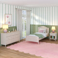 Pastel girls bedroom interior with twin bed, pink scalloped rug, green wainscoting, white dresser, bedside lamps, sheer curtains and scallop-pattern wallpaper