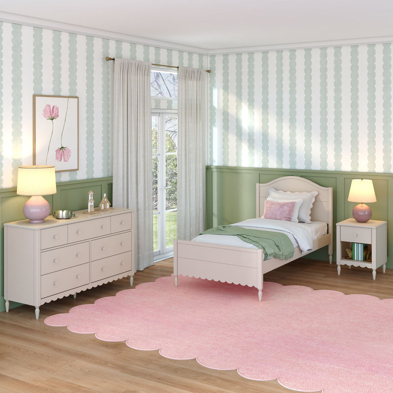 Pastel girls bedroom interior with twin bed, pink scalloped rug, green wainscoting, white dresser, bedside lamps, sheer curtains and scallop-pattern wallpaper