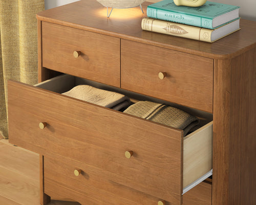 Mid-century modern wooden dresser with brass knobs and warm wood finish, open drawer with neatly folded clothes and a lamp and books on top.
