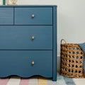 Blue painted dresser with brass knobs beside woven wicker basket and pastel striped rug in a nursery/kids’ bedroom