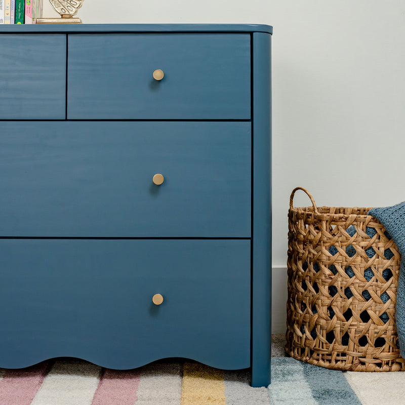 Blue painted dresser with brass knobs beside woven wicker basket and pastel striped rug in a nursery/kids’ bedroom