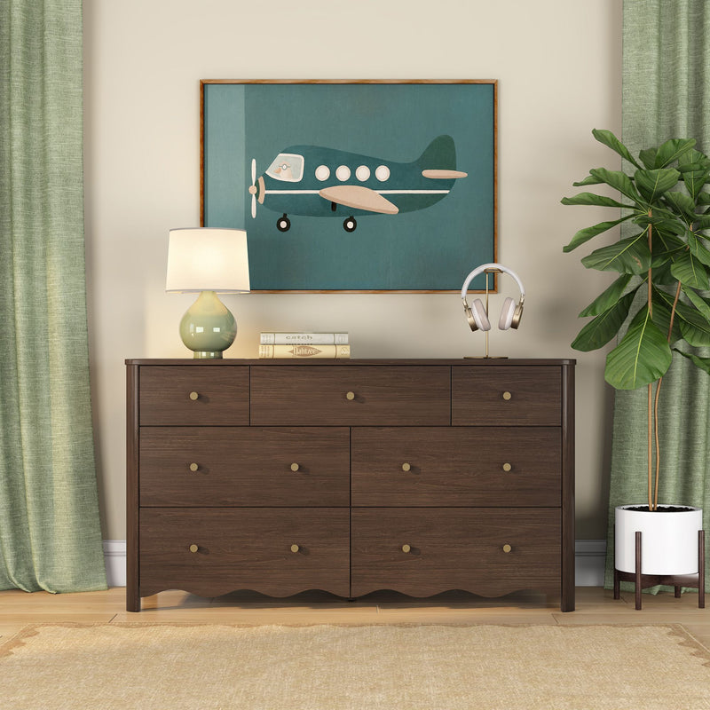 Dark walnut 6-drawer dresser styled in modern bedroom with teal airplane wall art, green ceramic table lamp, wireless headphones and fiddle leaf fig plant