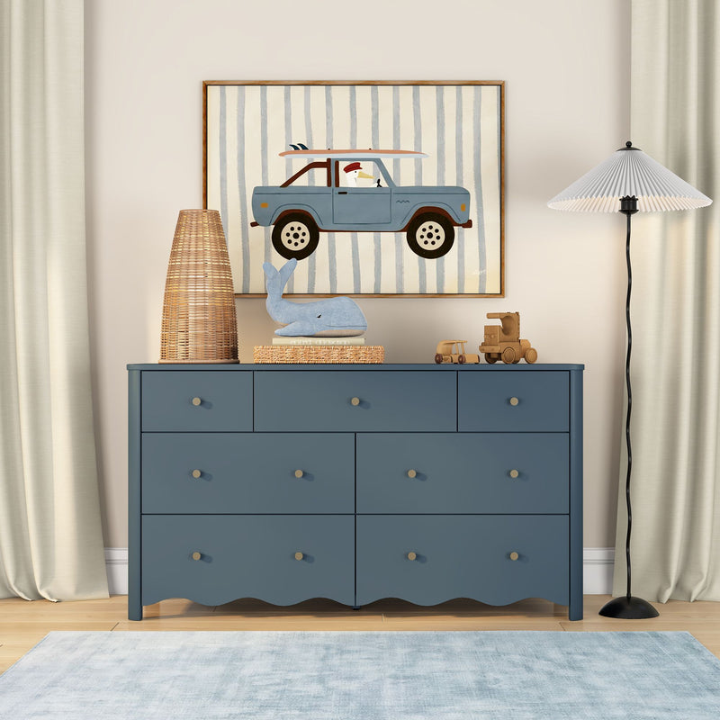 Coastal-themed nursery with blue dresser, framed car and surfboard wall art, rattan lamp, plush whale toy and wooden toy trucks