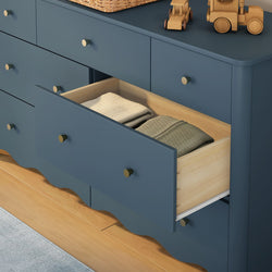 Navy blue wooden dresser in a nursery with an open drawer revealing folded baby clothes, brass knobs and wooden toy cars on a hardwood floor