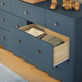 Navy blue wooden dresser in a nursery with an open drawer revealing folded baby clothes, brass knobs and wooden toy cars on a hardwood floor