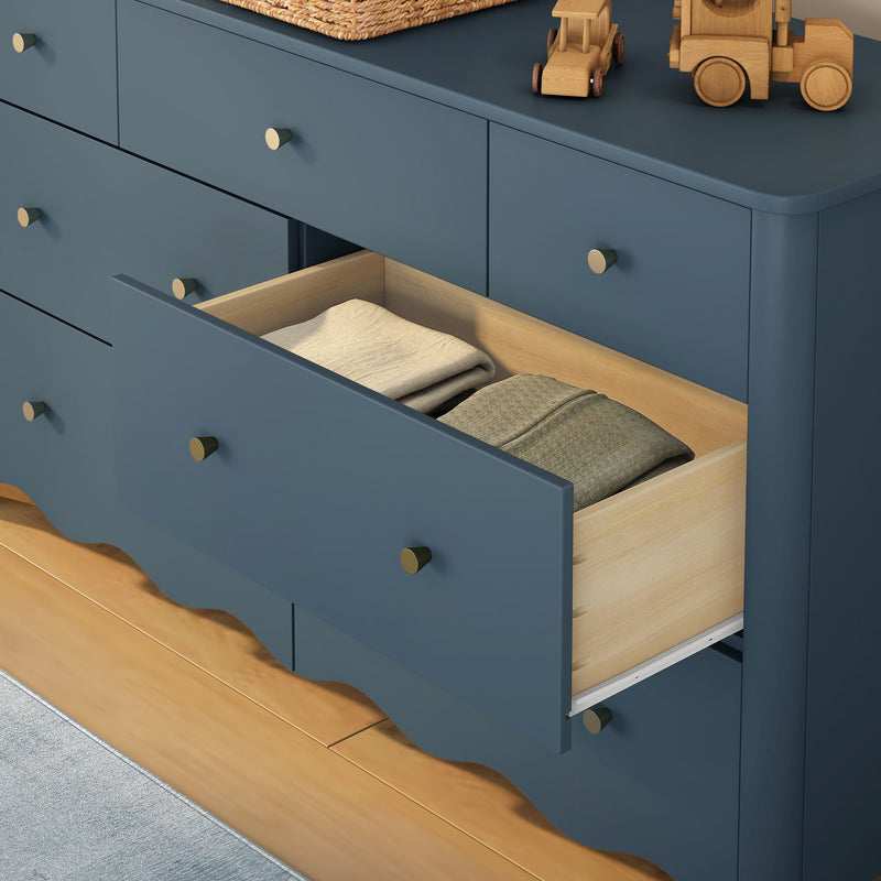 Navy blue wooden dresser in a nursery with an open drawer revealing folded baby clothes, brass knobs and wooden toy cars on a hardwood floor
