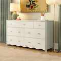 White nursery dresser with round brass knobs and scalloped base, styled with table lamp, wooden giraffe toy, colorful wall art, floor lamp and green curtains on hardwood floor