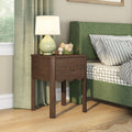 Walnut wooden bedside table with single drawer and ceramic lamp next to green upholstered bed with gray gingham bedding