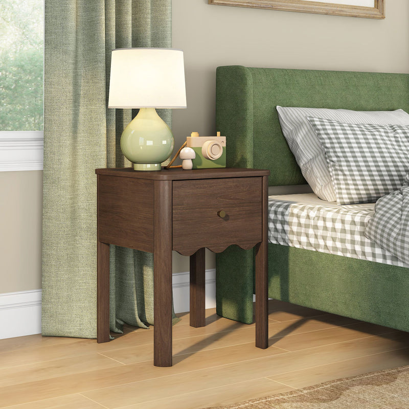 Walnut wooden bedside table with single drawer and ceramic lamp next to green upholstered bed with gray gingham bedding