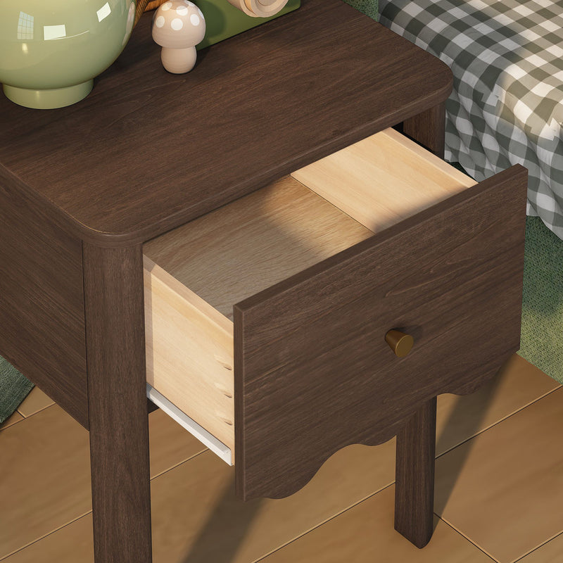 Walnut-finish wooden nightstand with open drawer and brass knob, scalloped apron and visible drawer joinery, green ceramic vase and wooden mushroom decor on top, gingham bedding and hardwood floor in cozy bedroom setting