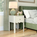White bedside table nightstand with drawer and green ceramic lamp next to green upholstered bed with gray gingham bedding and wooden toy camera