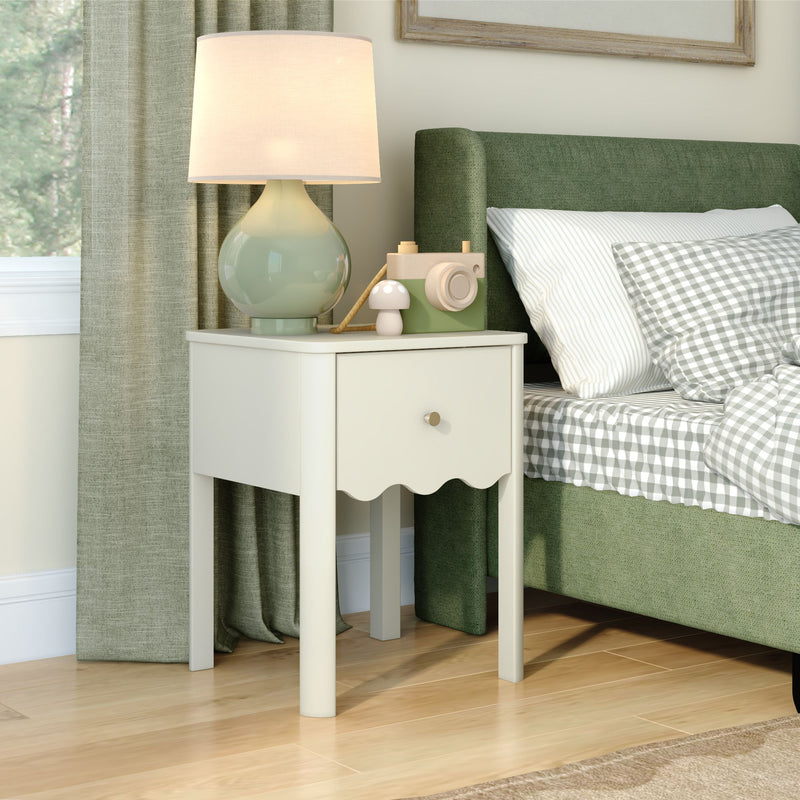 White bedside table nightstand with drawer and green ceramic lamp next to green upholstered bed with gray gingham bedding and wooden toy camera