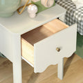 White bedside table nightstand with open light-wood drawer and brass knob, scalloped edge, lamp and mushroom decor on top beside checked bedding on hardwood floor