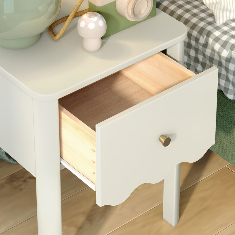 White bedside table nightstand with open light-wood drawer and brass knob, scalloped edge, lamp and mushroom decor on top beside checked bedding on hardwood floor