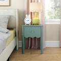 Teal bedside nightstand with scalloped drawer, yellow ceramic lamp and bunny alarm clock next to upholstered bed in cozy modern bedroom with pink curtains and natural light