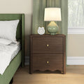 Dark wood two-drawer nightstand with brass knobs topped by a green ceramic table lamp and white alarm clock beside a green upholstered bed with striped bedding, green curtains and hardwood floor in a bright modern bedroom.