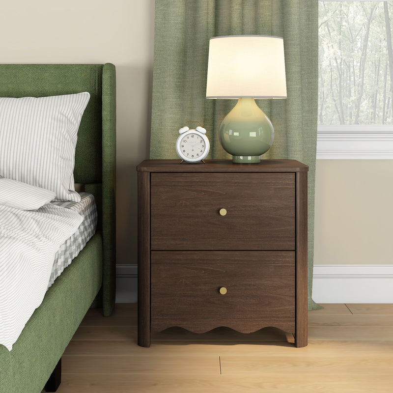 Dark wood two-drawer nightstand with brass knobs topped by a green ceramic table lamp and white alarm clock beside a green upholstered bed with striped bedding, green curtains and hardwood floor in a bright modern bedroom.