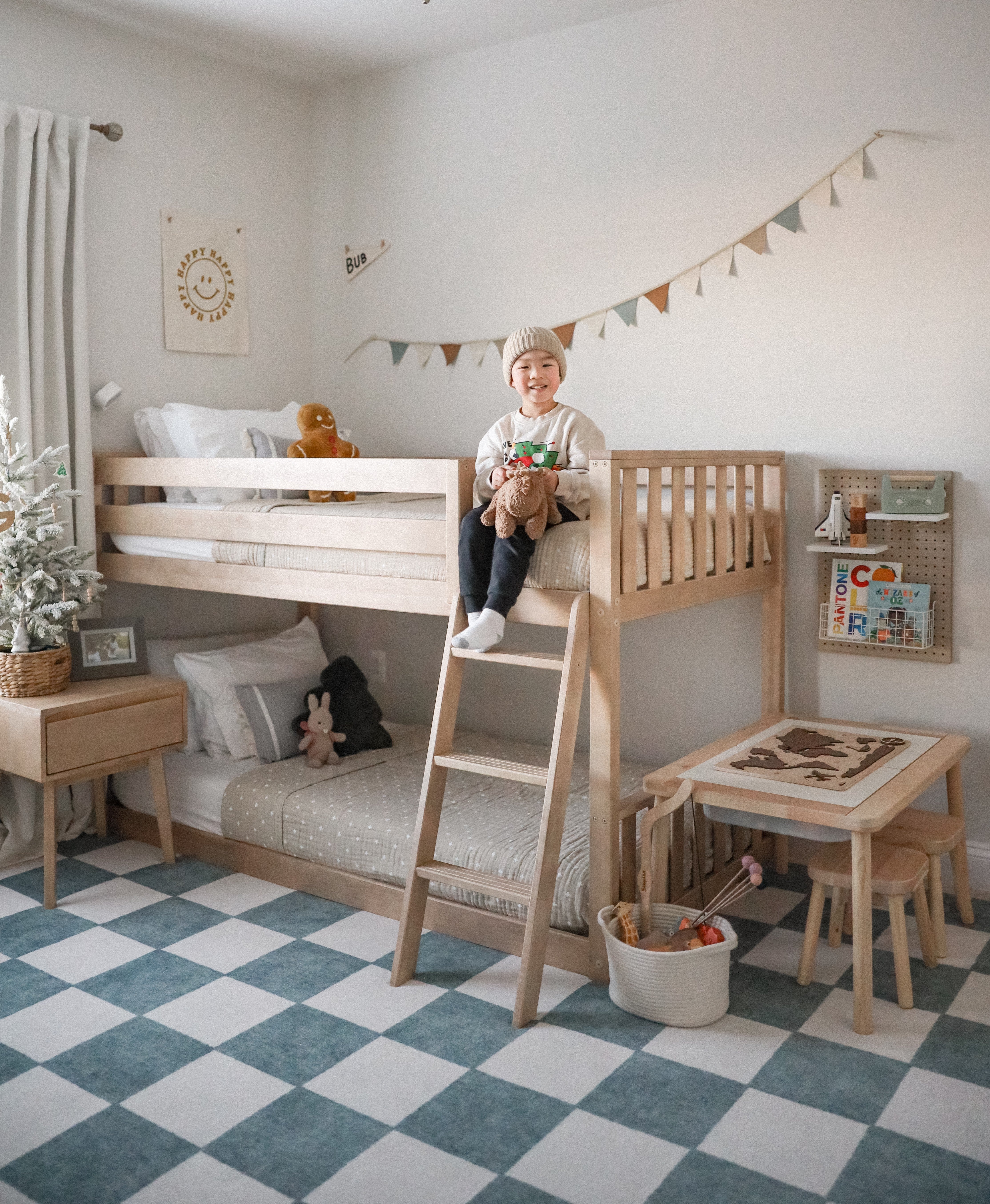 Young child sitting on a natural wood bunk bed in a cozy neutral Scandinavian-style kids' bedroom with a blue-and-white checkered rug, play table, stuffed animals and bunting.