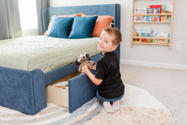 Child kneeling by blue upholstered bed pulling out under-bed storage drawer while holding a plush elephant toy in a bright kids bedroom with bookshelf