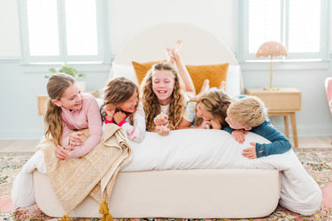 Five kids laughing on a cozy bed during a bright, family-friendly sleepover in a modern bedroom