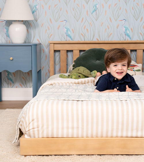 Smiling child on wooden toddler bed with striped bedding and plush turtle in coastal-themed kids' bedroom with heron-pattern wallpaper.