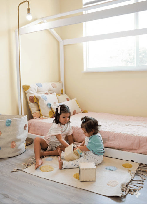 Two children playing on the floor of a bedroom with a bed and window in the background.