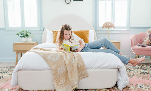 A young girl reading a book on white upholstered bed in a pink and blue room.