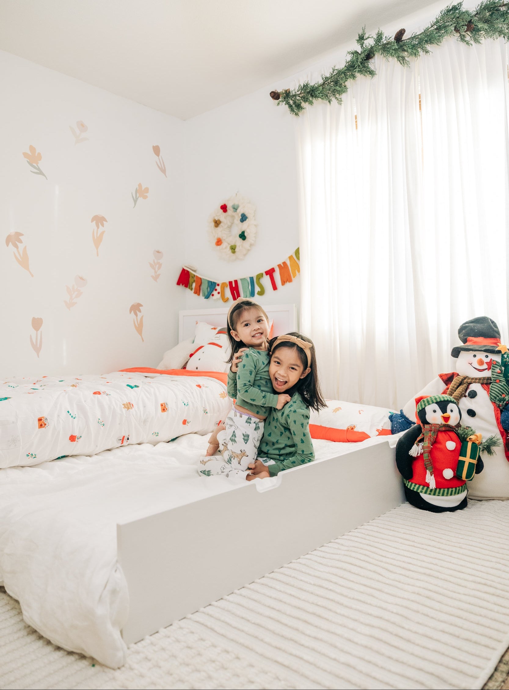 Two smiling children in matching green Christmas pajamas hugging on a white toddler bed in a bright, holiday-decorated kids’ bedroom with a “Merry Christmas” banner, garland, plush penguin and snowman toys, white curtains and floral wall decals