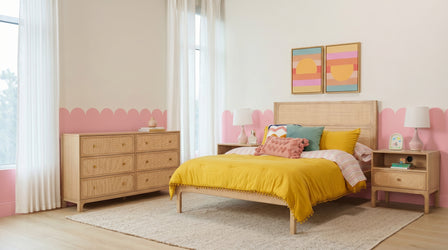 Modern pastel bedroom with scalloped pink wall trim, rattan bed and dresser, mustard yellow duvet, textured pillows, abstract sunset prints and sheer curtains