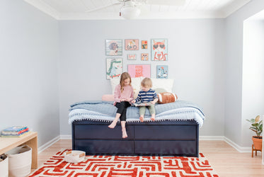 Two children reading on a navy storage bed in a bright modern kids' bedroom with pastel animal wall art, blue quilted bedding, and a red geometric rug
