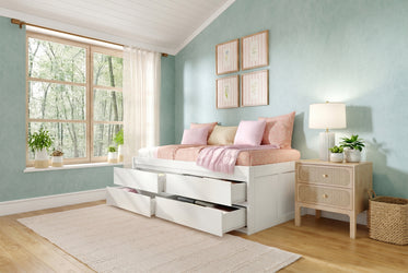 White storage bed with multiple under-bed drawers, pastel pink bedding and throw, mint green textured walls, large wood-frame window with sheer curtains, bedside nightstand, lamp and potted plants for small bedroom storage and decor ideas
